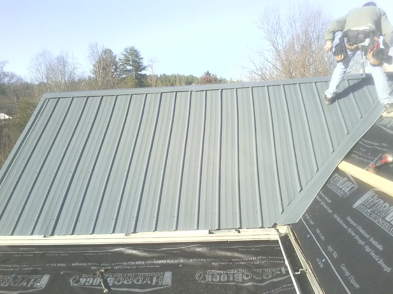 Crew installing dark green standing seam panels over synthetic underlayment in Manzanola