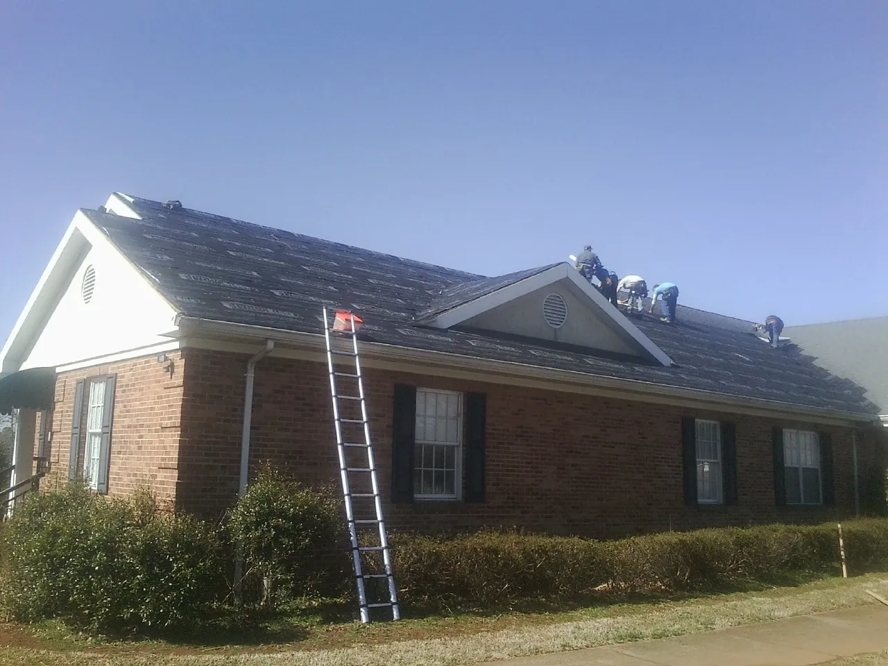 Crew tearing shingles off a brick ranch roof with a ladder set — Emergency Roof Repair work by our crew in Manzanola