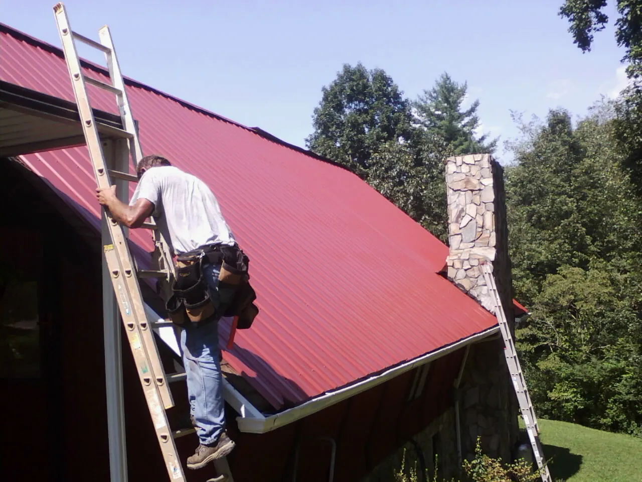 Roofer on a ladder beside a red metal roof and stone chimney — Soffit & Fascia Repair by Roofing Brothers in Manzanola